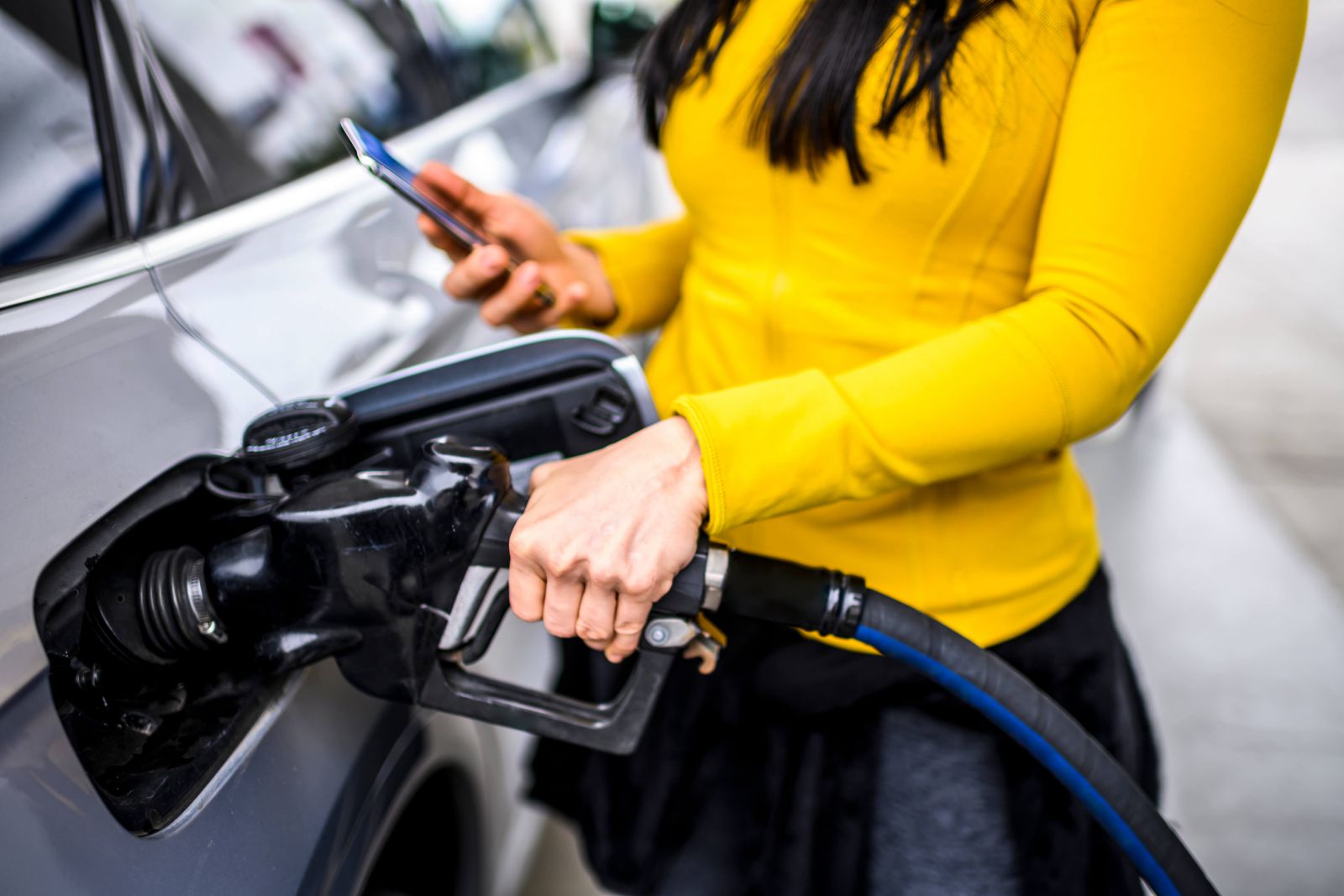 Person in yellow top using a smartphone while fueling a car with a black gas pump.